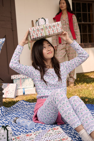 A child wears the SERFLEX Kids Snowday Stripe Iggy Play Set, featuring long sleeves and matching pants with a colorful horizontal stripe pattern of reindeer, snowflakes, and hearts.