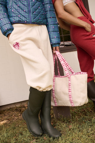 A person holds a beige sherpa Plaid Hearts Sherpa Tote with pink plaid handles and trim, coordinating with a matching pink plaid heart patch on their sweatpants.