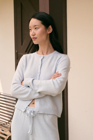 A woman poses with her arms crossed, wearing an icy blue AFFINA Moxie Cardigan with pearl-like buttons and matching drawstring pants.