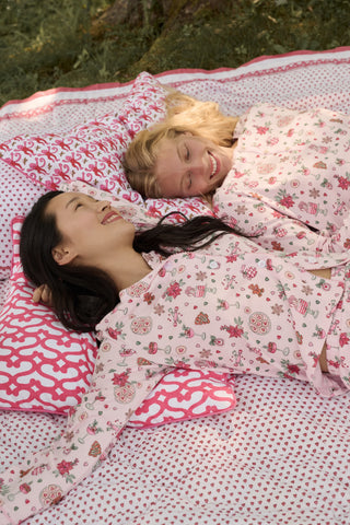Two women sit on a patterned blanket outdoors wearing matching light pink Seasons Soiree Hayden Pajamas by NETTALCO, featuring a classic Pima cotton top and ruffled shorts decorated with holiday-themed illustrations.