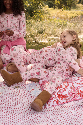 A child in a patterned Kids Seasons Soiree Juniper Pajamas two-piece set and rubber boots sits on a wooden sled and looks through binoculars.