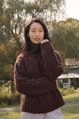 A person wearing a cozy, chocolate brown Naomi Sweater by REVOLUTION KNITS, featuring a cable-knit design and a high turtleneck.