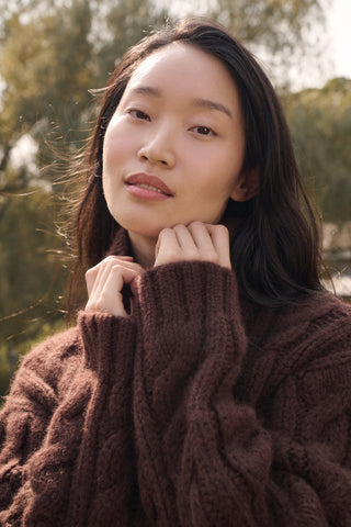 A person wearing a cozy, chocolate brown Naomi Sweater by REVOLUTION KNITS, featuring a cable-knit design and a high turtleneck.