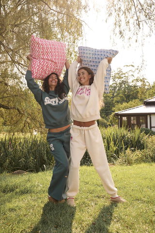 A woman wears the ARILCONFEX Sleep Club Andie Sweatshirt, an oatmeal crewneck with SLEEP in pink plaid appliqué across the chest and Club embroidered below it in pink script.