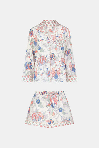 A woman stands against a plain white background, smiling slightly as she models the Amanda Leila Set from Roller Rabbit—a vintage-inspired cotton loungewear ensemble featuring a long-sleeve, button-up pajama top and matching shorts adorned with a colorful floral pattern.