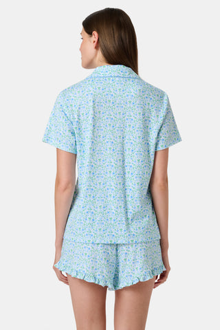 The Roller Rabbit Garden Gate Floral Wyatt Polo Pajamas feature a light blue short-sleeve set crafted from soft Pima Cotton with a delicate Garden Gate Floral Ditsy pattern. The button-up top is designed with a collar and chest pocket, while the matching shorts are accented with a charming ruffled hem. The background is plain white.