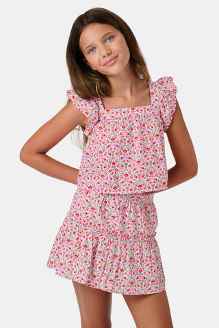 Three young girls sit close together outdoors, each wearing the Roller Rabbit Kids Garden Gate Floral Emmett Top—an ideal choice for girls' summer clothing. They are smiling and appear to be engaged in conversation, with greenery and stone walls in the background.