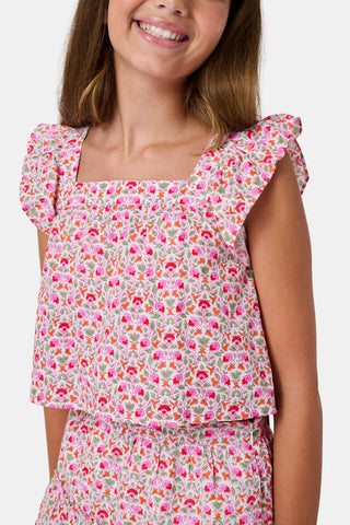 Three young girls sit close together outdoors, each wearing the Roller Rabbit Kids Garden Gate Floral Emmett Top—an ideal choice for girls' summer clothing. They are smiling and appear to be engaged in conversation, with greenery and stone walls in the background.