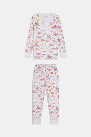 A young girl smiles barefoot in Roller Rabbit Kids Summer Memories Pajamas, made from soft Pima Cotton with playful, colorful prints on white. She stands against a light gray background. Made in Peru.