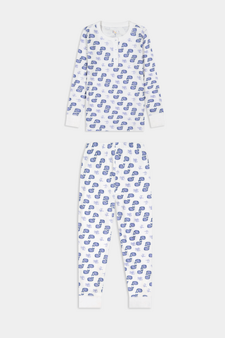 A young boy stands barefoot and smiling in Roller Rabbit's Kids Moby Pajamas—white Pima cotton pajamas with blue car prints, featuring long sleeves and pants—against a plain white background.