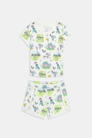 A girl stands outdoors in a Roller Rabbit Kids Game, Set, Monkey Playa Play Set, holding out a tennis ball toward the camera. Her playful outfit is perfect for kids’ tennis activities, and the photo is taken from a low angle with blue sky and trees in the background.