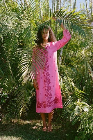 A person stands barefoot among lush tropical plants, partly hidden by palm fronds, wearing the bright pink Cross Stitch Vine Shiv Caftan from Roller Rabbit as sunlight filters through the leaves.