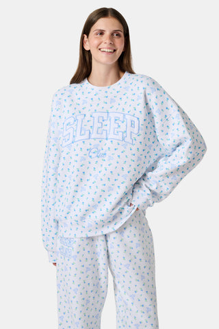 A person wearing a white Roller Rabbit Mini Monkey Blossom Andie Sweatshirt, featuring small blue and green Mini Monkey Blossom prints and large SLEEP Club lettering, stands outdoors, partially in sunlight. Their face is mostly out of frame.