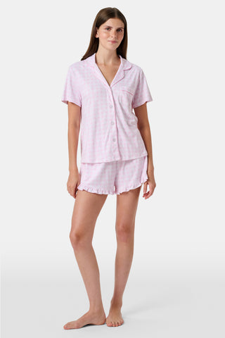 The Gingham Hearts Wyatt Polo Pajamas by Roller Rabbit are a pink and white gingham short-sleeve pajama set, featuring a button-up shirt with chest pocket and matching shorts with a ruffled hem, all crafted from soft Pima cotton. This set is shown on a white background.
