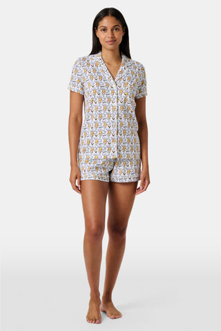 A woman stands barefoot, wearing the SERFLEX Vanderbilt Lovely Monkey Polo Pajamas—a matching short-sleeve button-up pajama top and shorts featuring a yellow and gray floral pattern on a white background.