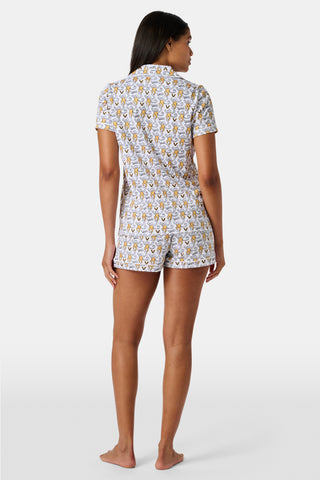 A woman stands barefoot, wearing the SERFLEX Vanderbilt Lovely Monkey Polo Pajamas—a matching short-sleeve button-up pajama top and shorts featuring a yellow and gray floral pattern on a white background.