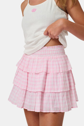 Three girls stand in front of lockers, holding notebooks. Smiling at each other, they are dressed in pink and white outfits, with one wearing the Roller Rabbit Kids Gingham Hearts Sami Skirt. Blue and pink backpacks are visible by the lockers.
