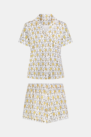 A woman stands barefoot, wearing the SERFLEX Vanderbilt Lovely Monkey Polo Pajamas—a matching short-sleeve button-up pajama top and shorts featuring a yellow and gray floral pattern on a white background.