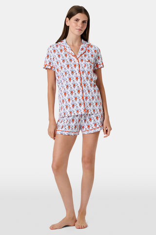 A woman stands barefoot against a plain white background, wearing the SERFLEX UVA Lovely Monkey Polo Pajamas—a matching short-sleeved top and shorts set featuring a festive red and white print with penguins and snowflakes reminiscent of school colors.
