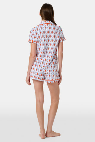 A woman stands barefoot against a plain white background, wearing the SERFLEX UVA Lovely Monkey Polo Pajamas—a matching short-sleeved top and shorts set featuring a festive red and white print with penguins and snowflakes reminiscent of school colors.