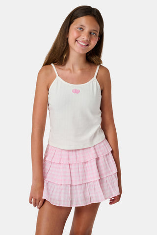The Kids Hearts Pointelle Bridget Cami by Roller Rabbit is a white sleeveless camisole with thin straps and a small pink heart-shaped patch at the center of the chest. Crafted from soft pointelle cotton, it features a delicate scoopneck design. The background is light gray.