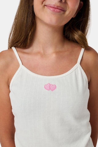 The Kids Hearts Pointelle Bridget Cami by Roller Rabbit is a white sleeveless camisole with thin straps and a small pink heart-shaped patch at the center of the chest. Crafted from soft pointelle cotton, it features a delicate scoopneck design. The background is light gray.