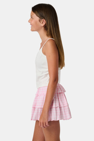 The Kids Hearts Pointelle Bridget Cami by Roller Rabbit is a white sleeveless camisole with thin straps and a small pink heart-shaped patch at the center of the chest. Crafted from soft pointelle cotton, it features a delicate scoopneck design. The background is light gray.
