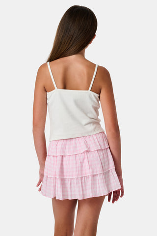 The Kids Hearts Pointelle Bridget Cami by Roller Rabbit is a white sleeveless camisole with thin straps and a small pink heart-shaped patch at the center of the chest. Crafted from soft pointelle cotton, it features a delicate scoopneck design. The background is light gray.