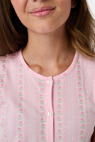 A young girl in a rose pink dress smiles while kneeling on the floor, holding a colorful folder. A pink backpack hangs nearby in a classroom or locker area, and she wears her cozy Roller Rabbit Kids Daisy Heart Stripe Diana Cardigan.