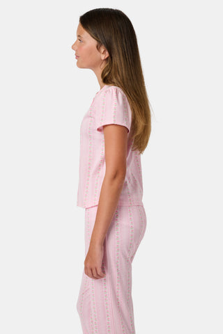 A young girl in a rose pink dress smiles while kneeling on the floor, holding a colorful folder. A pink backpack hangs nearby in a classroom or locker area, and she wears her cozy Roller Rabbit Kids Daisy Heart Stripe Diana Cardigan.