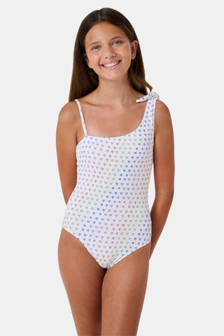 The Disco Hearts Kids Jacey One Piece by Roller Rabbit is a white swimsuit with rows of colorful hearts, featuring an asymmetrical neckline, one thin strap, and a knot-tie detail on the right shoulder.