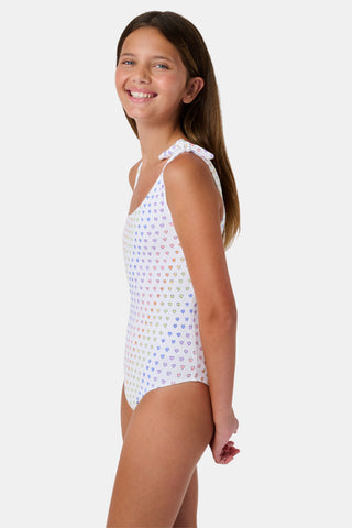 The Disco Hearts Kids Jacey One Piece by Roller Rabbit is a white swimsuit with rows of colorful hearts, featuring an asymmetrical neckline, one thin strap, and a knot-tie detail on the right shoulder.