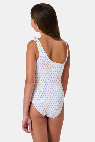 The Disco Hearts Kids Jacey One Piece by Roller Rabbit is a white swimsuit with rows of colorful hearts, featuring an asymmetrical neckline, one thin strap, and a knot-tie detail on the right shoulder.