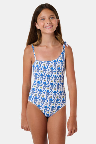 The Monkey Kids Jacey One Piece by Roller Rabbit is a white, one-shoulder swimsuit with blue monkeys and yellow bananas. It features an asymmetrical neckline, a knotted right tie strap, and playful monkey illustrations.