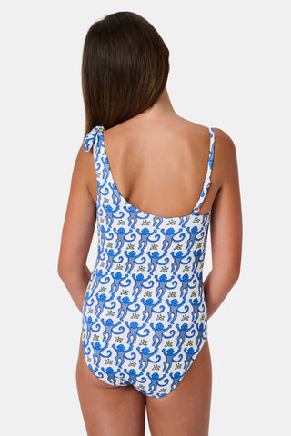 The Monkey Kids Jacey One Piece by Roller Rabbit is a white, one-shoulder swimsuit with blue monkeys and yellow bananas. It features an asymmetrical neckline, a knotted right tie strap, and playful monkey illustrations.