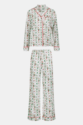 A woman wears a SERFLEX Deck The Halls Bailey Pajamas set featuring a short-sleeved top and shorts with a white background and a repeating Christmas-themed pattern of elves, snowmen, and evergreen trees.