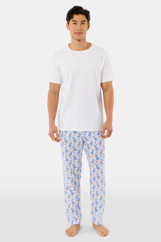 A person stands barefoot against a white background, wearing Roller Rabbit's Mens Romy the Rabbit Jon Pant—white vintage cotton pants with a blue and orange bunny print—one hand in their pocket, the other at their side.