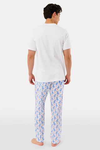 A person stands barefoot against a white background, wearing Roller Rabbit's Mens Romy the Rabbit Jon Pant—white vintage cotton pants with a blue and orange bunny print—one hand in their pocket, the other at their side.