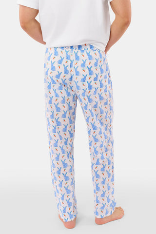 A person stands barefoot against a white background, wearing Roller Rabbit's Mens Romy the Rabbit Jon Pant—white vintage cotton pants with a blue and orange bunny print—one hand in their pocket, the other at their side.