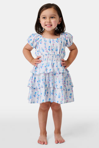 Four young girls stand and chat in front of white lockers, wearing pastel kids' dresses. One girl wears the Roller Rabbit Kids Naia Floral Stripe Alana Dress and holds a backpack and books, appearing to be in a school hallway. Another girl holds paper over her head.