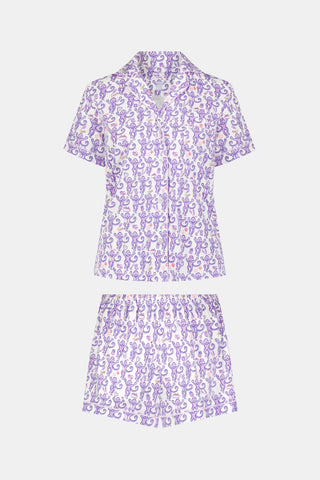 A woman is wearing the TEXPIMA Get Ready With Me Polo Pajamas — a matching short-sleeve button-up top and shorts set featuring a purple and pink patterned print — while standing against a plain light background, looking at the camera with a gentle smile.