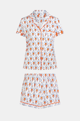 A woman stands barefoot against a plain white background, wearing the SERFLEX UVA Lovely Monkey Polo Pajamas—a matching short-sleeved top and shorts set featuring a festive red and white print with penguins and snowflakes reminiscent of school colors.