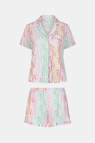 A woman with curly hair, standing and smiling slightly, wears Roller Rabbit’s Disco Monkey Wyatt Polo Pajamas: a short-sleeve button-up top and matching ruffled shorts featuring a pastel multicolor abstract pattern on white.