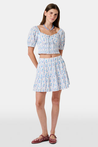 Four young girls stand and chat in front of white lockers, wearing pastel kids' dresses. One girl wears the Roller Rabbit Kids Naia Floral Stripe Alana Dress and holds a backpack and books, appearing to be in a school hallway. Another girl holds paper over her head.