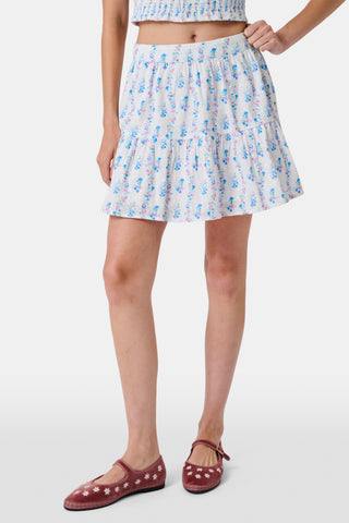 Four young girls stand and chat in front of white lockers, wearing pastel kids' dresses. One girl wears the Roller Rabbit Kids Naia Floral Stripe Alana Dress and holds a backpack and books, appearing to be in a school hallway. Another girl holds paper over her head.
