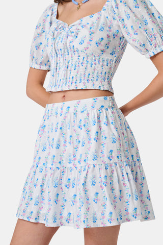 Four young girls stand and chat in front of white lockers, wearing pastel kids' dresses. One girl wears the Roller Rabbit Kids Naia Floral Stripe Alana Dress and holds a backpack and books, appearing to be in a school hallway. Another girl holds paper over her head.