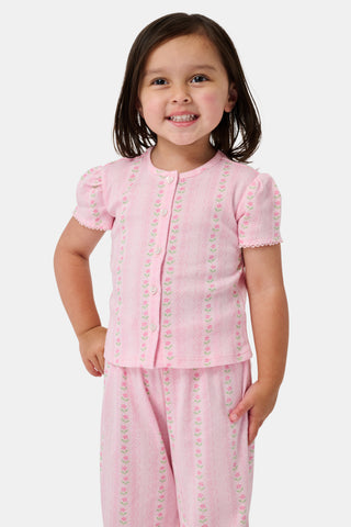 A young girl in a rose pink dress smiles while kneeling on the floor, holding a colorful folder. A pink backpack hangs nearby in a classroom or locker area, and she wears her cozy Roller Rabbit Kids Daisy Heart Stripe Diana Cardigan.