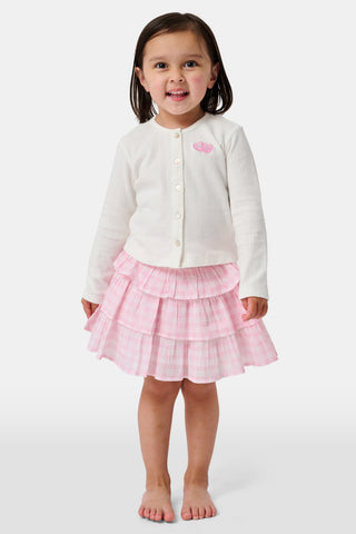 Three girls stand in front of lockers, holding notebooks. Smiling at each other, they are dressed in pink and white outfits, with one wearing the Roller Rabbit Kids Gingham Hearts Sami Skirt. Blue and pink backpacks are visible by the lockers.