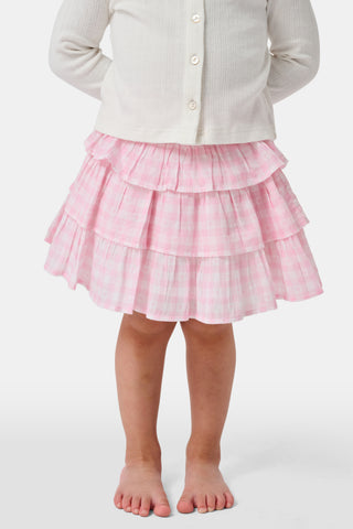 Three girls stand in front of lockers, holding notebooks. Smiling at each other, they are dressed in pink and white outfits, with one wearing the Roller Rabbit Kids Gingham Hearts Sami Skirt. Blue and pink backpacks are visible by the lockers.