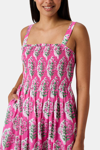 The Kids Ava Floral Ceri Dress by Roller Rabbit is a sleeveless baby dress featuring a smocked bodice and ruffled straps, adorned with green and white leaf patterns on a vibrant pink background, and comes with matching bloomers.
